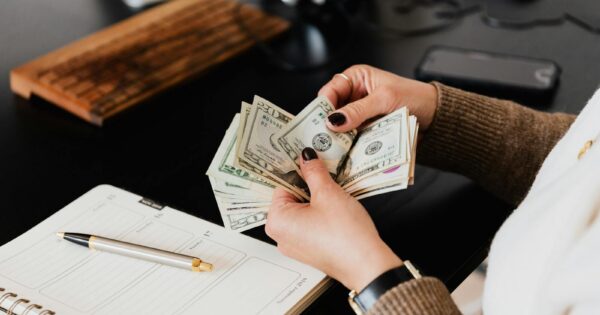 Unrecognizable elegant female in sweater counting dollar bills while sitting at wooden table with planner and pen